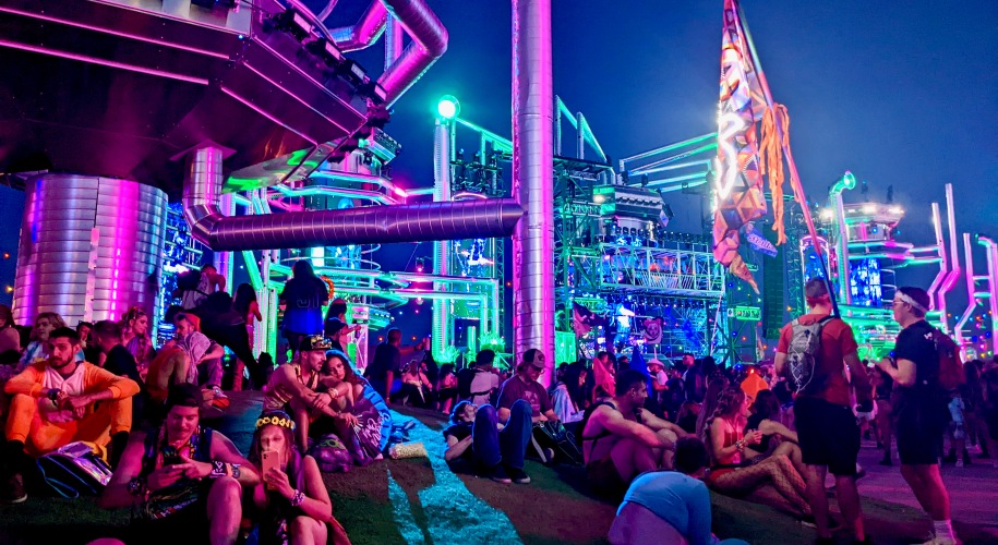 men and women sitting on the ground underneath an elaborate metal stage structure at nighttime, EDM electronic music festival rave setting with neon colored lights